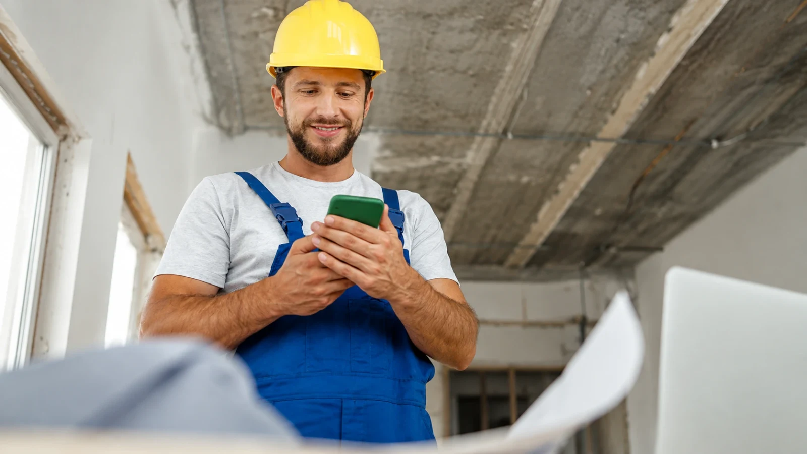 Construction worker in a hard hat logging his hours on a phone at an unfinished site