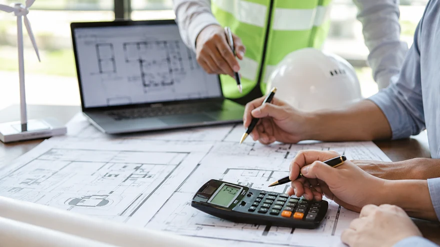 Site team reviewing project costs on blueprints with a calculator and laptop